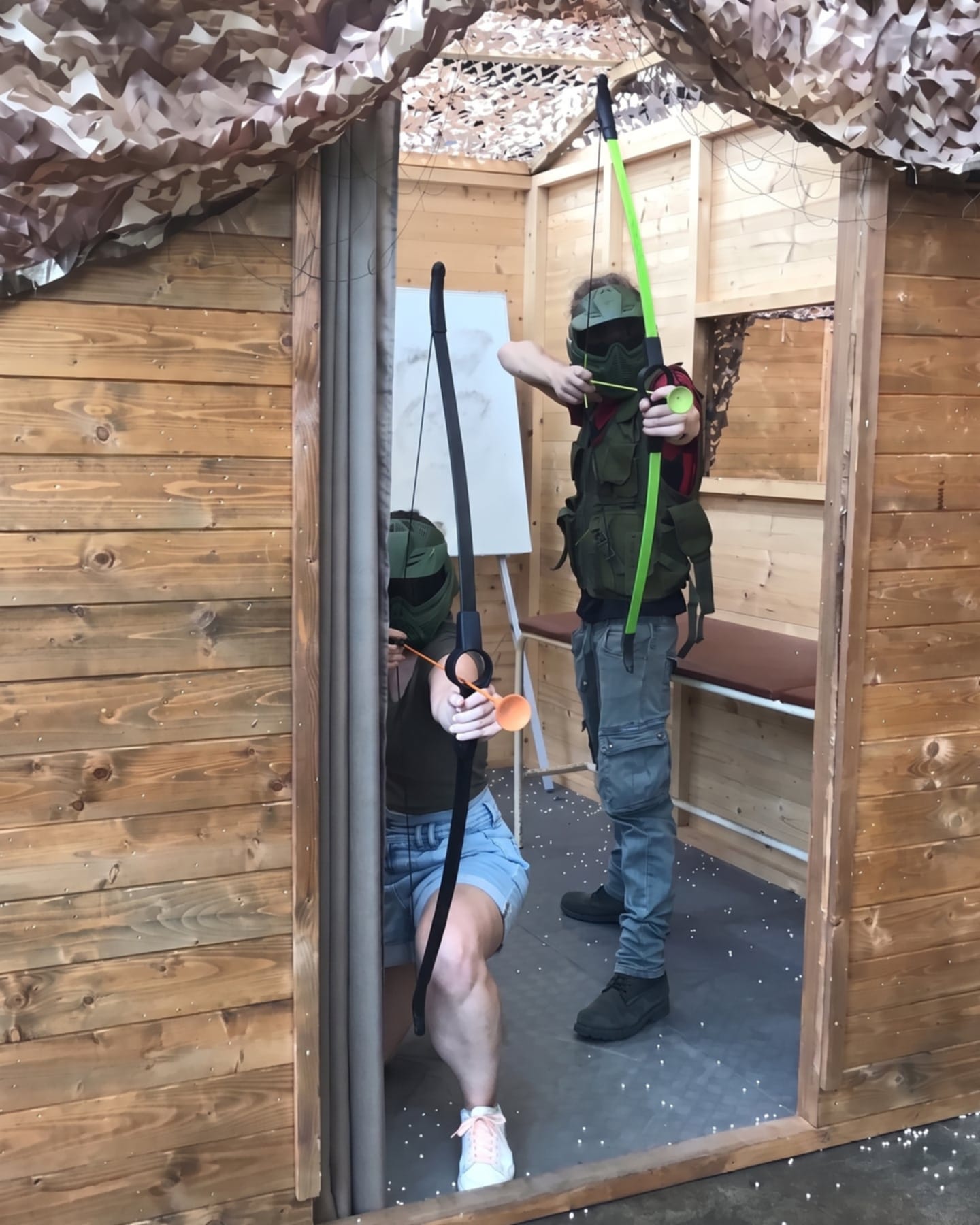 Due arcieri con maschera protettiva mirano con l'arco dalla porta di una casetta di legno coperta da una rete mimetica.