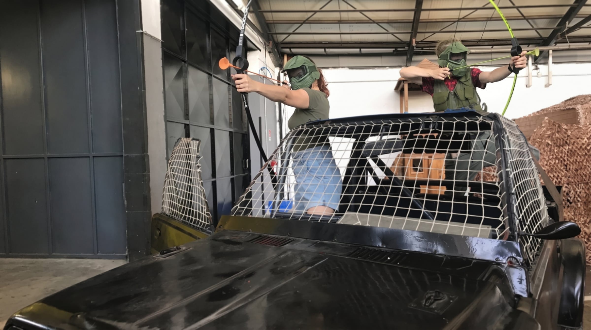 Due giocatori di arcotag con maschera protettiva tendono l'arco sopra una jeep con la rete, nell'arena indoor di Softair Milano.