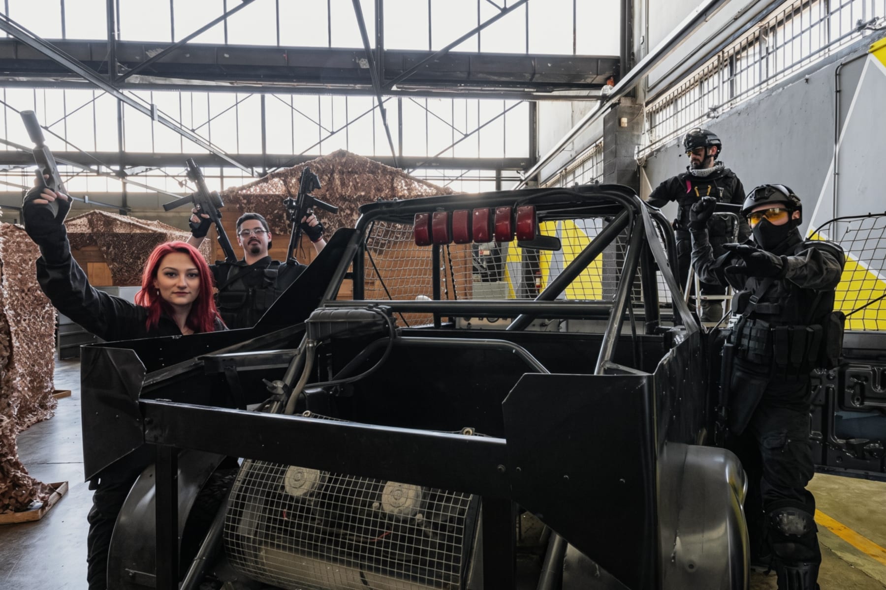 Il capannone dell'arena al coperto: un buggy con la gabbia e giocatori in tenuta tattica sotto le capriate e i lucernari.