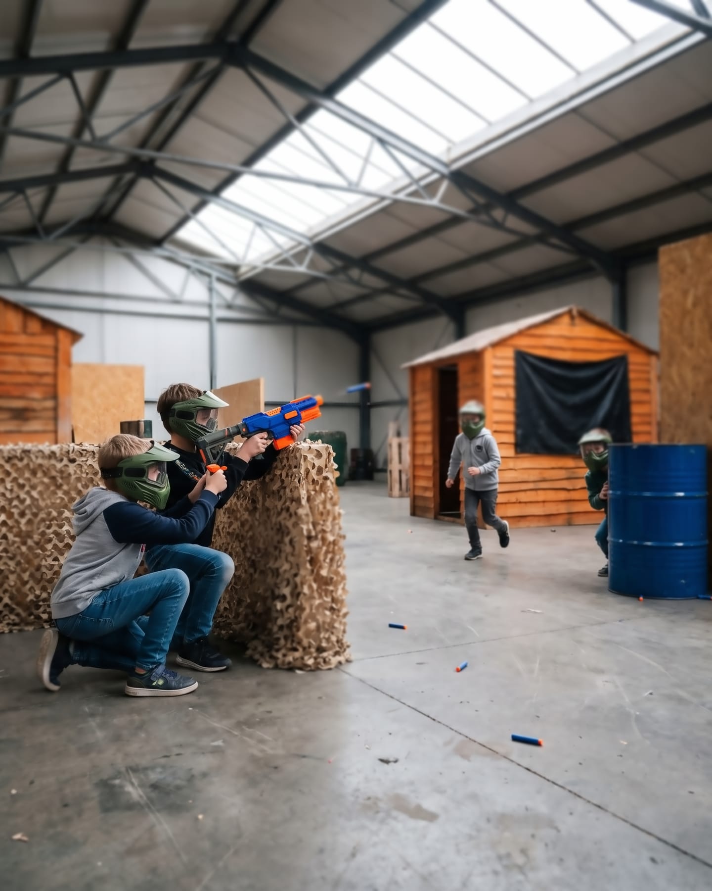 Bambini con maschera protettiva e blaster Nerf tra casette di legno e reti mimetiche: due, accovacciati dietro una barricata, sparano un dardo in gommapiuma, un terzo corre e un quarto sbuca da dietro un bidone blu.